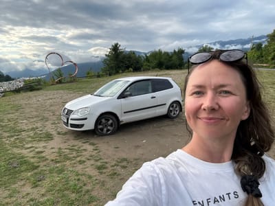 A person is taking a selfie outdoors, with a white car parked behind them. The person is wearing a white t-shirt with black text and sunglasses. Their hair is pulled back with a black scrunchie. The car appears to be a white minivan. The background shows a grassy area, dirt, trees, mountains, and a cloudy sky. The image is oriented sideways, with the person's head at the top and feet towards the bottom. The text on the t-shirt reads "LES ENFANTS". There is also a heart-shaped metal sculpture visible in the background. The time of day appears to be overcast, likely late afternoon or early morning, given the soft light. The location is Adzhakhy, Abkhazia. The overall mood seems relaxed and casual.