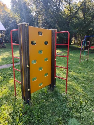 The image depicts a playground scene on a sunny day. In the foreground, a yellow climbing wall with rectangular cutouts is supported by dark wooden posts and framed by red metal bars. Yellow balls are visible on the climbing wall and its supports. To the right of the climbing wall, a partially visible ladder-like structure with red supports and green and blue rungs stands in the grass. The background features a grassy area with trees and sunlight filtering through the leaves. To the left, a portion of a white-roofed structure, possibly a gazebo or shelter, is visible. The ground is covered in green grass and some weeds. The overall setting suggests a recreational area in an outdoor environment. This scene is likely located in or near the city of Катунь, Russia, given the context. There are no people present in the image.