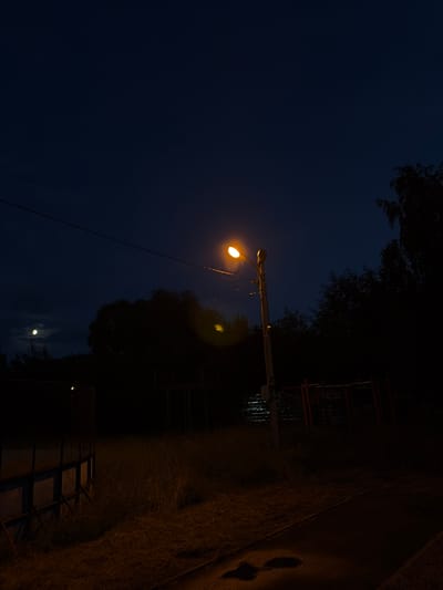 The image depicts a night scene in Martem'yanovo, Russia. A single streetlamp, illuminated with a warm yellow light, is the most prominent object in the foreground, casting a glow on the surrounding darkness. The lamp is mounted on a pole and is positioned to the left of the frame, extending horizontally.
The background is dominated by the deep blue of the night sky, with some trees silhouetted against it. A faint glow from the moon is visible in the lower part of the sky, partially obscured by clouds. To the right of the streetlamp, there are some structures that appear to be part of a playground or recreational area, with red horizontal bars and vertical supports.
The ground in the right portion of the image is paved, possibly a walkway or road, and shows some reflections, suggesting dampness or recent rain. Patches of grass and dry vegetation are visible along the edge of the paved area and extending into the darker regions of the scene. The overall mood of the image is tranquil and somewhat solitary, characteristic of a quiet night in a suburban or rural setting. There is no visible text in the image.