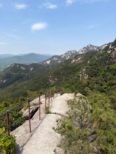 Captured in Goyang-si, South Korea, the image presents a precarious mountain trail. A pathway of large, light-colored rocks is visible, bordered by a rudimentary safety railing of metal poles and cables. Beyond the trail, a steep, verdant slope descends into the hazy distance, revealing the tops of successive mountain ridges under a light blue sky with a few scattered clouds. The perspective gives the impression of height and exposure.