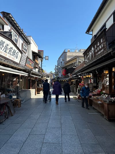 The image shows a narrow street lined with buildings and shops in Katsushika City, Japan (35.758, 139.877). Four people are walking along the street, all wearing face masks. Three people are wearing dark clothing, and one is wearing a purple jacket. The street is paved with gray square tiles, and the buildings are mostly made of wood. There is a sign above the street with Japanese writing. There are shops on both sides of the street, selling various items. The sky is clear blue.
