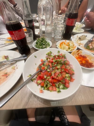 A table setting shows a variety of dishes. A large white bowl holds a chopped salad of tomatoes, cucumbers, and red onion. Other dishes contain chopped parsley, cabbage salad, hummus, and tomato sauce. Two glass bottles of Coca-Cola are lying on their sides near several empty drinking glasses. Silver serving utensils are strewn across the table, and the edge of a black handbag is visible on the right.