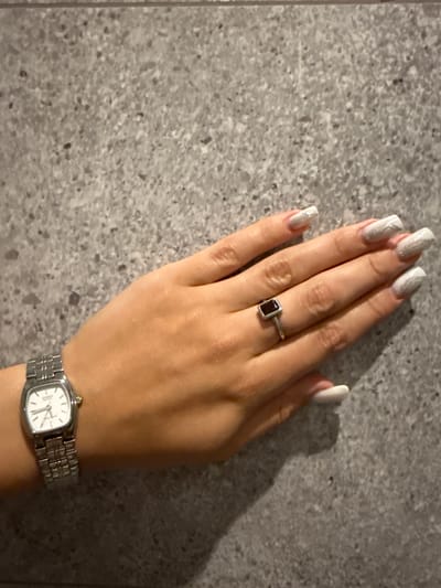 A left hand, adorned with white, glittery acrylic nails and a silver ring with a dark rectangular stone, rests on a textured grey surface. A silver, rectangular-faced wristwatch with a metal band is worn on the wrist. The background is a mottled grey surface, possibly a wall or countertop. The image is taken from a high angle, looking down at the hand and wrist. There is no identifiable location in the city of Pavlohrad, Ukraine.