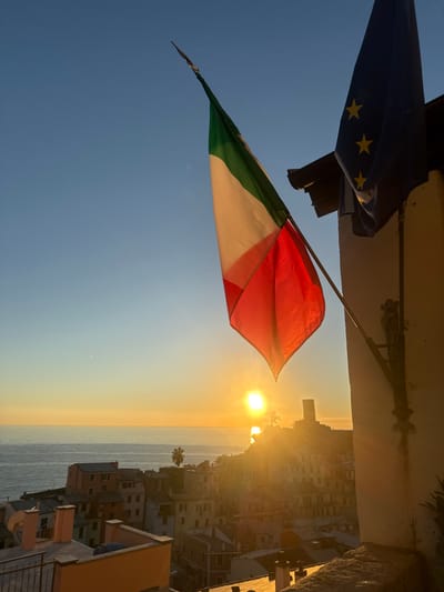 This image captures a high-angle view of Vernazza, Italy, at either sunrise or sunset, looking west over the Ligurian Sea. In the upper right quadrant, an Italian flag, displaying vertical green, white, and red stripes, is prominently mounted on a light-colored building. Adjacent to it, a European Union flag, depicting a blue field with twelve yellow stars, is also mounted but largely obscured in shadow. A dark eave extends from the building above the flags.
The sun is visible as an intensely bright disc low on the horizon, casting a strong golden glare that reflects on the calm surface of the sea. The sky transitions from a clear, light blue overhead to a warm orange and yellow hue near the horizon.
Below the flags, the colorful, clustered buildings of Vernazza cascade down the hillside towards the coastline. These structures are illuminated by the warm, direct light of the low sun. On the distant right, a distinct stone tower, characteristic of Doria Castle, stands silhouetted against the sun's glare. Several rooftops and a balcony with an iron railing are visible in the immediate foreground. Sparse vegetation, including palm trees, is interspersed among the buildings and on the distant hills. No human figures or specific activities are depicted.