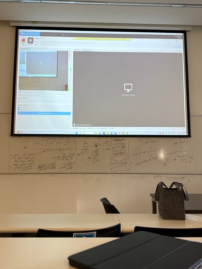 A classroom in Kfar Monash, Israel, located at 32.342 latitude and 34.911 longitude, features a projector screen displaying a paused screen recording of a lecture. Handwritten equations and diagrams cover the whiteboard. A black laptop lies on a desk in the foreground, while a gray tote bag sits on a separate desk to the right. Two black chairs are visible in the background. The scene suggests an academic setting.