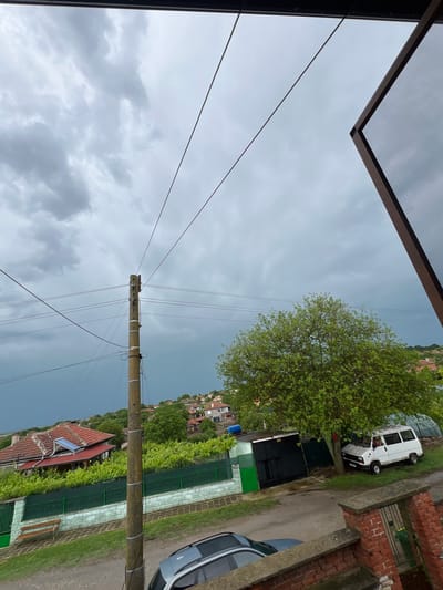 The image depicts a rural or village street scene in Ботево, Bulgaria, under an overcast sky. The overall impression is one of a quiet, somewhat rustic residential area on a cloudy day, potentially before or after a rain shower.
In the foreground, the upper left corner shows the dark frame and mesh of what appears to be a window or screen, hinting that the photo was taken from inside a building looking out. Several thin power lines extend from the top of the frame, traversing the sky.
A prominent concrete utility pole stands upright in the midground, heavily laden with multiple overhead power lines that stretch across the frame, connecting to unseen points. Some white markings, too small to decipher, are visible on the pole.
To the right of the pole and in the midground, a dirt or gravel road is visible. A white, older model utility van is parked facing left on the grass verge beside this road. Closer to the viewer on the right, the front portion of a dark-colored car is partially visible, parked by a low brick wall. Along the road and behind the vehicles, there are green metal fences and lush green foliage, possibly hedges or vines. Several sheds or small structures, some green and some dark, are also present.
Further into the background, a cluster of houses with distinctive red-tiled roofs can be seen, nestled among green trees. One house in the closer background features a visible solar panel on its roof. The landscape includes distant hills or rising terrain, also dotted with houses and greenery. A large, leafy green tree stands tall near the white van.
No people are visible, and there is no discernible activity taking the foreground, midground, or background. The heavy, grey clouds suggest dim daylight, possibly early morning or late afternoon, and create a muted atmosphere.