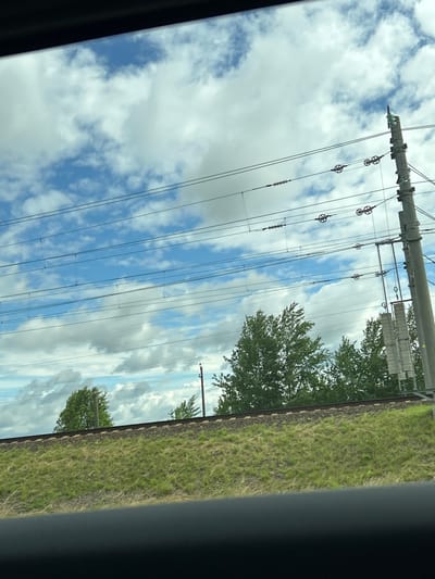 The image captures a view from a moving vehicle, looking out over a grassy embankment with railway tracks. Above the embankment, a line of trees is visible against a partly cloudy blue sky. Several parallel overhead power lines, supported by a utility pole on the right, stretch across the frame. The scene suggests a rural or semi-rural setting, possibly on the outskirts of a settlement like Малевіцкі сельскі Савет, Belarus. There are no people or animals visible in the frame. The overall impression is one of a journey through a landscape dominated by infrastructure and natural elements.
