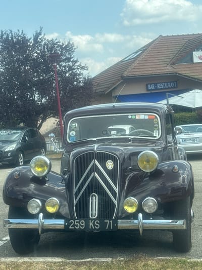 The image shows a vintage dark purple Citroën Traction Avant parked in front of a building with a "BAR - RESTAURANT" sign. A dark grey Peugeot hatchback is parked to the left of the Citroën, and a silver convertible with license plate "AR 405 WK" is visible to the right. The scene appears to be outdoors on a sunny day with scattered clouds. The Citroën has a license plate reading "259 KS 71". The building has a tiled roof and a blue awning. The foreground shows grass and pavement. The image is cropped to focus on the front of the vintage car.