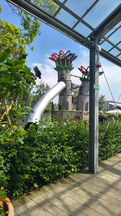 The image shows a whimsical, treehouse-themed playground with a large silver slide curving out from a wooden structure resembling a tree trunk. The playground features colorful, petal-shaped decorations at the top of the structures. In the foreground, a lush green hedge lines a paved walkway. A dark grey metal pole stands to the right of the hedge, supporting parts of a glass-roofed structure overhead. A man in a black shirt is visible in the background, facing away from the camera, near a white umbrella and a building. The scene is set outdoors with a bright blue sky and green trees visible in the background. The location is identified as a playground in Rain, Germany.