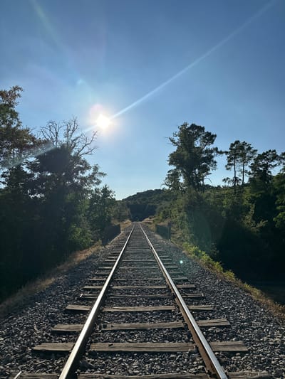 The image shows a railway track leading towards a tunnel in a rural area near Générargues, France. The sun is shining brightly, creating lens flare in the sky and illuminating the scene. Trees and vegetation line the sides of the tracks, which are laid on a bed of ballast. The sky is clear blue. The scene is peaceful and natural, with no people or animals visible.