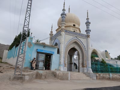 The image depicts a mosque in Bostan, Pakistan. The mosque features a large central dome flanked by smaller minarets. An ornate arched gateway leads to the mosque entrance. Adjacent to the mosque is a small blue building with a storefront. A man in traditional Pakistani clothing is leaning against the building. A power line pole is on the left. A modern-looking building is in the background. A blue metal fence encloses part of the mosque grounds. The sky is overcast.