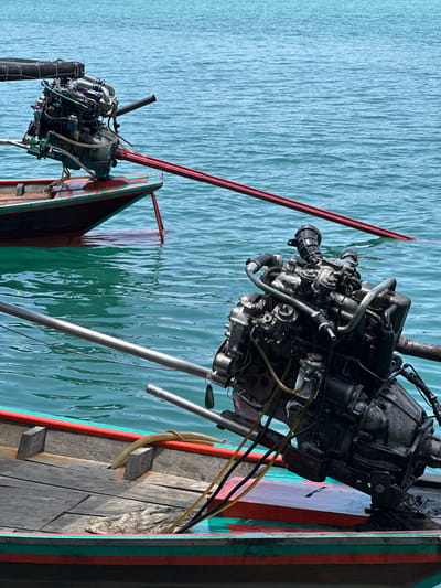 The image captures two traditional Thai longtail boats, each equipped with a converted car engine mounted high on a pivoting frame. These engines are positioned above the waterline, with the propeller shaft extending downward into the water. The boats are situated on the surface of the placid Cheow Lan Lake, a body of water located in Ban Ta Khun, Thailand. The lake's surface reflects the ambient light, creating a shimmering effect. The boats' wooden hulls show signs of wear and use, with peeling paint and exposed wood grain. The engines are complex assemblies of metal components, hoses, and wires, showing evidence of modification and adaptation. The boats are likely used for transportation, fishing, or tourism activities on the lake.