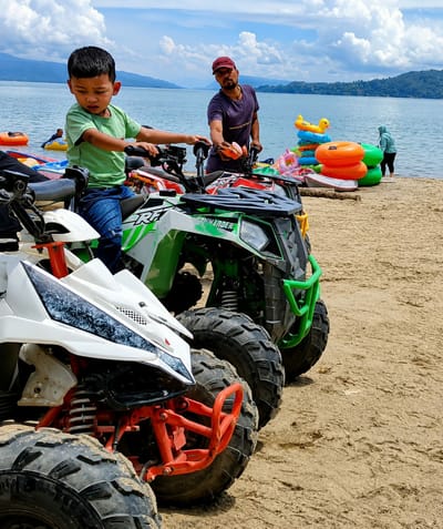 The image depicts a beach scene in Lumban Bul Bul, Indonesia, with several all-terrain vehicles (ATVs) lined up on the sandy shore. In the foreground, two ATVs are prominently visible. The one on the left is white with red accents and large black tires. The ATV to its right is green and black, also with substantial tires. A young boy in a green t-shirt and blue jeans is seated on the green ATV, appearing to be in control of the handlebars. Behind him, a man wearing a purple t-shirt and a red cap stands next to another ATV, holding an orange object. In the background, a large body of water, likely a lake, stretches towards a treed hillside under a cloudy sky. Several inflatable tubes, including a yellow duck and orange rings, are stacked on the beach near the water's edge. A woman in a light blue hijab is visible near the water. The overall atmosphere suggests a recreational setting.