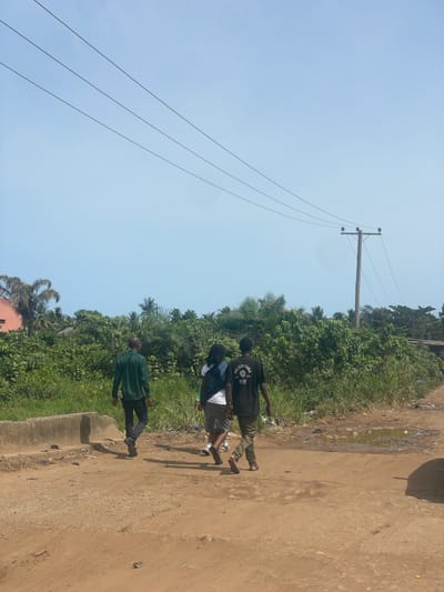 The image depicts a scene in Ikoga, Nigeria. Three individuals are walking along a dirt road bordered by dense vegetation. An electrical pole and power lines are visible against a bright blue sky. The road appears unpaved and somewhat uneven. The individuals are dressed in casual attire. The surrounding vegetation is lush and green, suggesting a tropical climate. There is some scattered debris along the roadside.