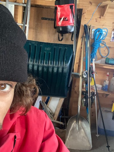 The image depicts a close-up indoor scene, likely a storage area or shed, in La Salle-les-Alpes, France. In the foreground, partially visible on the left, is the upper-left quadrant of a person's face and upper torso. The person has medium brown skin, dark brown eyes, and brown wavy hair visible beneath a black knitted beanie. They are wearing a red jacket with a visible zipper pull on the chest pocket. The person is looking directly forward towards the camera.
The background consists of wooden plank walls, suggesting an rustic indoor structure. Various items are stored within the space. A large, dark green plastic snow pusher with vertical ribs is prominently positioned vertically in the center-left. Above it, a red and black "PETZL BUCKET 15L" bag is hanging from a metal support bar.
To the right of the snow pusher, a pair of blue and white alpine skis with bindings are leaning against the wall, alongside two ski poles. A metal shovel with a wooden handle stands upright on the floor near the skis. Further right, a coiled blue rope hangs on the wooden wall, above a wooden shelf holding various bottles and containers, some blue and white. A section of a metal ladder is visible leaning against the wall behind the snow pusher. The overall impression is a cluttered but organized utility space for outdoor and winter gear.