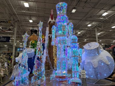 Inside a store, Christmas decorations are displayed. A large, iridescent nutcracker statue dominates the view, adorned with colorful lights. Flanking it are a Christmas tree-shaped decoration with a star and a large ornament, also shimmering with lights. In the background, a traditional nutcracker stands behind a slender, lit decoration, adding depth to the holiday scene.