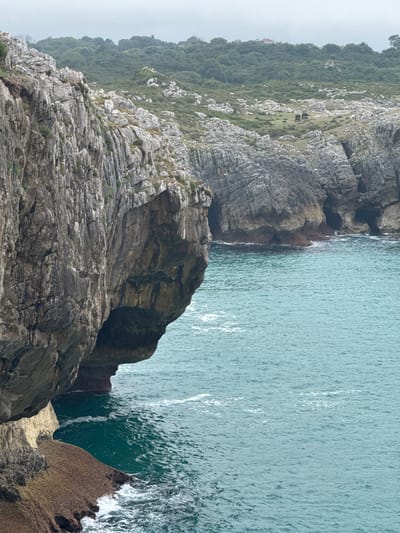 The image depicts a rugged coastal landscape with imposing cliffs overlooking a turquoise sea. In the distance, a green, hilly terrain is visible, dotted with trees and a few small structures, suggesting a sparsely populated area. The cliffs themselves are weathered and layered, with visible erosion and dark openings that appear to be caves or sea arches. The water exhibits ripples and small waves, indicating some movement. The overall scene is natural and serene, with the dramatic cliffs contrasting with the calm expanse of the ocean and the verdant hills in the background. The location appears to be along the coast of Llanes, Spain, a region known for its striking cliffs and natural beauty.
