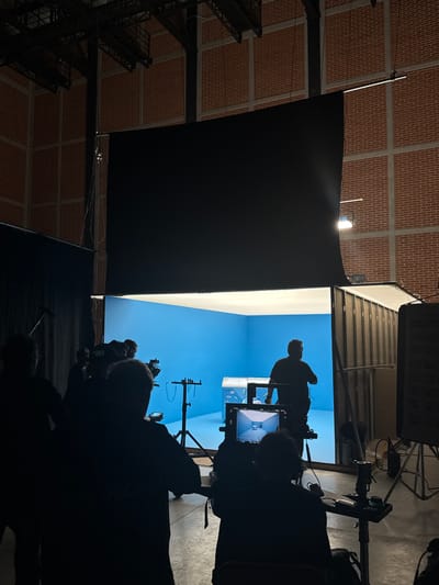 The image shows a film or video set located inside a building with a brick ceiling. A blue screen backdrop is visible. Several people are present, but they are mostly silhouetted. One person stands on a platform in front of the blue screen. Filming equipment, including cameras and monitors, are pointed towards the subject. The location is likely Tlalnepantla, Mexico.