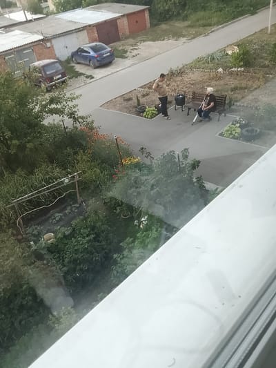 The image depicts an outdoor scene, likely in a residential area of Krasnoarmeysk, Russia. In the foreground, a reflective surface, possibly a window, obscures part of the view.
The main subjects are two people and several objects. A man in a light-colored t-shirt and dark pants stands to the left, facing a woman seated on a park bench. The woman, with white hair, is dressed in dark clothing and holds a walking cane. A black trash can is positioned behind the man. Behind the bench, a small garden area with plants and landscaping elements is visible. In the background, there are garages with cars parked in front of them, including a blue sedan and a dark-colored hatchback. A paved walkway or road runs between the garages and the garden area.
The scene appears to be set during the daytime, likely late afternoon or early evening, given the soft lighting and shadows. The weather seems to be pleasant, with no indication of rain or strong winds. The presence of garages and residential buildings suggests an urban or suburban environment.
There are no overtly discernible emotions, but the interaction between the man and woman could suggest a conversation or brief encounter. The overall impression is one of everyday life in a neighborhood. No visible text is apparent on any of the objects or structures.