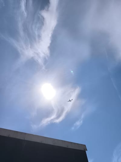 The image shows a clear blue sky with wispy white clouds. The sun is visible, casting a bright glare. An airplane is flying in the distance. In the foreground, there is a dark, flat surface with a concrete edge, possibly the roof of a building. The perspective suggests this is a view looking upwards towards the sky from a structure in Uithoorn, Netherlands.