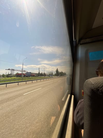The image captures a view from inside a vehicle, likely a bus or train, looking out a window. The perspective is unusual, as the image is rotated approximately 90 degrees clockwise.

In the foreground, the interior of the vehicle is visible. A dark-colored seat with a textured surface is on the right side, and a portion of a person's dark clothing is also visible. Above, a light-colored panel lines the interior of the vehicle.

The main scene observed through the window is a highway with multiple lanes. The road surface is asphalt, and white lane markings are clearly visible. On the left side of the highway, there is a clear blue sky with scattered white clouds, suggesting fair weather. The sun is shining, casting lens flare across the left side of the frame, indicating it's likely daytime, possibly mid-morning or afternoon.

Along the right side of the road, there are green trees and vegetation. In the distance, there are several structures and signs. One prominent blue sign with white text, which appears to be in Cyrillic, reads "ЮГ-КОВКА" (YUG-KOVKA) and includes a phone number. There are also some industrial-looking structures, possibly cranes or scaffolding, visible in the background. The scene suggests a rural or suburban setting along a major road. The overall atmosphere is one of travel and transit.