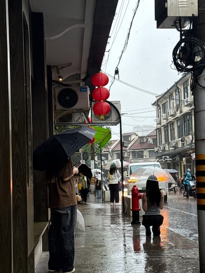 This is a vertical, eye-level, medium shot photograph taken on a rainy day on a street in Xuhui District, China. In the foreground, a person stands under a large black umbrella, facing away from the camera and looking down at their phone. Their legs are visible, clad in dark trousers and white shoes. To the right, another person is crouching on the wet sidewalk, holding a clear umbrella over their head, also facing away from the camera. A red fire hydrant stands to their left.
The sidewalk is wet and reflects the surrounding lights and structures. A white vehicle is parked in the street behind the crouching person, with its hazard lights on. Further down the street, a person on a blue scooter is visible, wearing a helmet and a blue outfit. Buildings with multiple windows line the street on the right side of the image. Red lanterns hang from a pole on the left side of the frame, near an air conditioning unit mounted on the building. The sky is overcast, and raindrops are visible in the air. The overall scene depicts a typical urban street during a rain shower, with people seeking shelter under umbrellas and navigating the wet conditions.