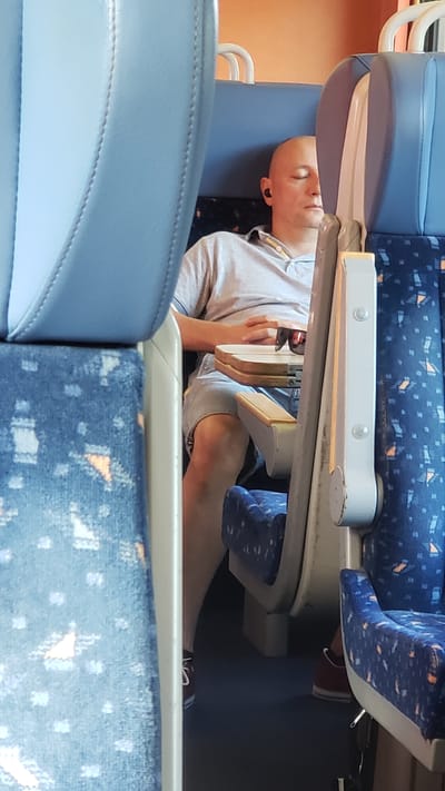 A bald man is seated on a train, sleeping with his eyes closed. He is wearing a light grey polo shirt and grey shorts, with a pair of black earbuds in his ears. A pair of sunglasses rests on a small foldable table in front of him. The seats are upholstered in blue fabric with a repeating pattern. The background shows the interior of a train car. There is no information available about the city of Považská Bystrica, Slovakia in relation to this image.