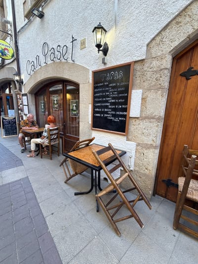 This media file captures a narrow street scene in Tossa de Mar, Spain, likely during daytime, with an overcast sky suggested by the lighting. The focal point is the entrance to a restaurant named "Can Paset." The building features a textured white stucco facade with a large, arched entryway leading to a wooden door and windows. A black chalkboard menu, displaying "TAPAS" at the top, lists various Spanish dishes such as "ENSALADILLA RUSA," "PATATAS BRAVAS," and "GAMBAS AL AJILLO."
Two individuals are seated at an outdoor table, appearing relaxed as they dine or converse. One person, with red hair, is looking down at the table, while the other, an older man with a patterned shirt, is leaning forward, also engaged with something on the table. Their presence suggests a leisurely atmosphere.
Several objects contribute to the scene's character: a vintage-style black lantern hangs from the stucco ceiling, a wooden ladder is propped against the wall near the entrance, and various chairs and tables are set up outside. A sign for "KARAOKE" and World Cup match times is also visible. The cobblestone and tiled pavement further ground the location as a Mediterranean street. The overall impression is one of a charming, established local eatery on a quiet street.