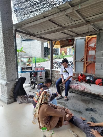 The image shows a scene in Kecamatan Purwoasri, Indonesia, located at coordinates -7.635 latitude and 112.136 longitude. It depicts a group of people sitting under a covered awning in front of a building. There is a food cart labeled "Bakwan" to the left, and a motorcycle parked behind it.  A man in a white shirt and blue jeans sits on a wooden bench to the right, while another man wearing a blue shirt and dark pants sits next to him.  A woman wearing a black headscarf sits on the ground to the left. There is a woman wearing a brown dress and a headscarf sitting next to the woman in black. A man wearing grey pants and a dark shirt lies on the ground, with his legs resting on the woman in brown’s lap.  He is massaging the woman in brown’s feet with one hand, while holding a cigarette in the other. There are a few helmets scattered on the ground. The scene takes place in a residential setting, with a concrete wall and a corrugated roof.