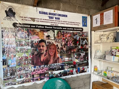 The image displays an indoor scene within a functional building, likely a community or aid center in Nzulo, Democratic Republic of the Congo. A large informational banner is prominently displayed on a rustic wall, while a tall, narrow shelving unit filled with supplies stands to its right.

The banner is a welcome and informational display for "GLOBAL BRIGHT CHILDREN "GBC" Asbl" and "The Kivu Project." It announces "THE IMPLEMENTATION OF HUMANITARIAN ACTIONS TO ASSIST VULNERABLE PEOPLE IN THE EAST OF THE DEMOCRATIC REPUBLIC OF CONGO, NORTH KIVU PROVINCE YEAR 2023." The banner explicitly welcomes "Father Daniel Torsiello and Jackie Jaramillo," whose smiling portraits are centrally featured. A collage of numerous smaller photos surrounds their images, depicting various humanitarian activities: distribution of bags of food and other aid, community gatherings, and groups of children and adults participating in programs, often outdoors. These images underscore the aid efforts of the organizations.

To the right, a simple, multi-tiered wooden shelf unit contains various packaged goods. The top shelf holds a cardboard box labeled "HEAVY DUTY LARGE" with a large '1' sticker. Text on the box indicates, "All items in this box contain commercial donations paid for and imported by The Kivu Project GBC asbl not recognized by the United States of America's government." The shelves below are neatly stocked with white packages, boxes of medication, blister packs, and bandages, suggesting a supply of medical or hygiene products for distribution. The overall scene depicts a hub for humanitarian operations, highlighting support for vulnerable populations in the specified region.