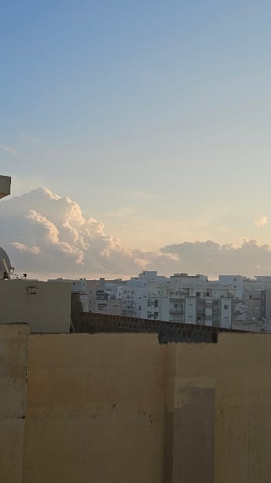 The scene depicts a bright, expansive sky transitioning from blue to a light peach color, filled with fluffy, sunlit clouds. A dense urban landscape with multi-story white buildings sits to the right of the clouds. In the foreground, a weathered, tan-colored structure with visible layers of concrete and aging texture provides a sense of depth and perspective. The composition is vertically oriented.