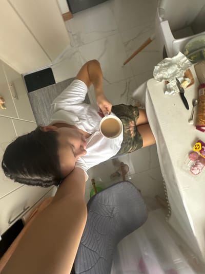 The scene is an interior view within a domestic kitchen or dining area in Gribovo, Russia. An individual with dark hair, wearing a white t-shirt and camouflage shorts, is seated at a white table. The person is holding a white mug filled with a light brown liquid in their right hand. The nails on the right hand are painted light grey. On the white tabletop, covered partially by a white cloth, are a partially unwrapped stick of butter on a small wooden cutting board, a black-handled knife, a silver spoon, an orange cylindrical container, and a clear plastic package containing three pink lollipops with a yellow "M" logo. The floor is covered with white tiles featuring grey veining. A grey textured mat is positioned under the seated individual. A section of a white high chair is visible to the right of the person. In the lower left quadrant, a dark grey textured cushion or mat is visible, partially obscured by the individual's left arm. White kitchen cabinets with silver handles are visible on the far left. A dark rectangular object rests beneath the cabinets. Further back on the tiled floor, two light-colored slippers and a wooden stick are present.