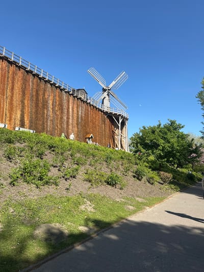 This image captures a large wooden graduation tower (Gradierwerk) under a clear blue sky, likely in Bad Rothenfelde, Germany. The structure, which dominates the left side of the frame, features a prominent four-bladed windmill at its upper section. The tower exhibits a dark, reddish-brown, textured appearance, characteristic of brine trickling over bundles of blackthorn brushwood, with visible wooden scaffolding and support beams. A long wooden ladder or walkway is positioned along the top edge of the tower.
In the upper left quadrant of the vibrant blue sky, a faint, crescent moon is visible, indicating a daytime shot. To the right of the graduation tower, a paved path or sidewalk runs parallel to the structure. This path is bordered by areas of green grass and low-lying bushes on both sides, with some patches of bare earth. Several trees with dense green foliage are scattered along the landscape, particularly behind the tower and adjacent to the path.
Shadows of people are distinctly cast onto the paved path, suggesting individuals are walking or standing nearby. Two small, light-colored figures, presumed to be people, are subtly visible in the mid-ground, standing near the base of the graduation tower. The scene is brightly lit by the sun, casting clear and defined shadows, indicating a sunny day. No visible text is present in the image.