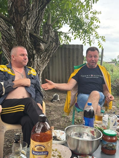 In Komsomolskiy, Russia, two men are seated outdoors under a large tree, possibly in a backyard or a rural setting. The man on the left, wearing a dark blue and yellow jacket over a bare chest and black pants with yellow stripes, sits on a light-colored plastic chair, looking to his right. He has a silver chain with a cross pendant. To his right, another man, wearing a black Tommy Hilfiger t-shirt and blue denim shorts, is seated on a green plastic chair. He is wearing a yellow jacket draped over his shoulders and appears to be speaking or reacting to something, with his mouth open and his left hand extended outwards. Between them, a small, grey and white kitten is visible on the ground. A table in front of them is laden with various items, including bottles of alcohol, a large metal bowl, plates with food, a jar of what appears to be pickled vegetables, and a bottle of Pepsi. The background shows a corrugated fence and vegetation. The lighting suggests it is daytime, with an overcast sky.