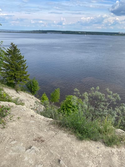 The image displays a panoramic daytime view captured from a high vantage point, likely a cliff or bluff, overlooking a vast body of water. The sky occupies the upper portion of the frame, a clear blue adorned with scattered, fluffy white clouds.

Below the sky, a wide expanse of dark blue-gray water stretches horizontally across the middle ground. The water's surface is textured with numerous subtle ripples, suggesting a gentle breeze. On this body of water, a small white sailboat is visible in the distance, indicating recreational activity or passage.

The far bank across the water is a long, low-lying strip of land, densely covered with green trees and vegetation. Faintly visible along this distant shoreline are some structures, possibly buildings or industrial elements, and a prominent, long bridge-like structure with multiple spans extending across the water.

The foreground is dominated by the steep, rocky, and dirt-covered edge of the cliff from which the photo was taken. This cliff face is sparsely covered with various types of green vegetation, including small bushes, brighter green deciduous trees, and darker green coniferous trees. In the lower right corner of the foreground, some small flowering plants with pinkish-purple blossoms add a touch of color to the otherwise earthy tones. A small, black circular object, possibly a discarded cap or lid, is visible on the dirt near the cliff's edge.

No people or visible text are present in the image. The scene, set in a location like "Пятый километр, Russia," evokes a sense of vastness and natural tranquility under a bright, partly cloudy sky.