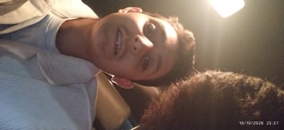A close-up, rotated photograph depicts a young male positioned from a low angle in a dimly lit, interior environment within the city of Al-Kiswa, Syria. The male subject, exhibiting a light complexion and dark hair, is smiling, revealing visible dental braces on his upper teeth. He is wearing a light-colored, possibly light blue, collared shirt with a textured pattern. His head is tilted to the right relative to the frame. A dark, textured mass, consistent with human hair or animal fur, is visible in the lower right quadrant, partially illuminated. A bright, rectangular light source is present in the upper right, casting significant glare and serving as the primary illumination. The background is dark and indistinct. A timestamp "18/10/2025 22:27" is overlaid in the lower right corner.