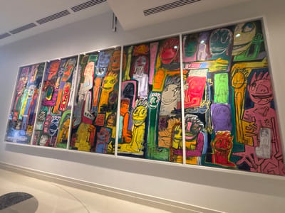 A large, multi-panel artwork is displayed prominently on a white wall within an indoor public space in Winchester, United States of America. The artwork is composed of at least seven vertical panels, each encased in a white frame, collectively featuring numerous highly stylized, brightly colored humanoid figures and faces. These figures are outlined in black and occupy the entirety of the canvas space, set against a dark background. The surrounding environment includes plain white walls, a white ceiling with multiple linear air vents, and a polished, light-colored floor exhibiting a partial dark circular geometric motif. The scene is brightly illuminated, with reflections visible on the artwork's surface. No individuals are present within the captured scene.