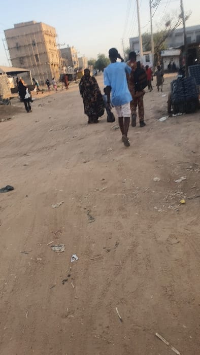 A street scene is captured on a sandy terrain. Buildings and utility poles line the left side, while the ground is strewn with trash. Three figures are visible: one in a blue shirt, another with a floral patterned dress, and a third nearby. More people are seen in the background near the buildings. A stack of square containers sits on the edge of the left side street, adding to the urban environment.