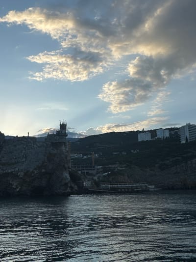 The image depicts the Swallow's Nest castle, a historical landmark located in Гаспринский поселковый совет, Ukraine. The castle is perched on a rocky cliff overlooking the Black Sea, with the water body occupying the right side of the frame. The horizon is partially obscured by the cliff on which the castle sits and the mountainous terrain to the left. The sky, occupying the top half of the image, is filled with clouds, and the lighting suggests either sunrise or sunset. The architecture of the castle is visible against the sky. The image appears to be taken at an angle, possibly from a boat or another vantage point at sea level.
