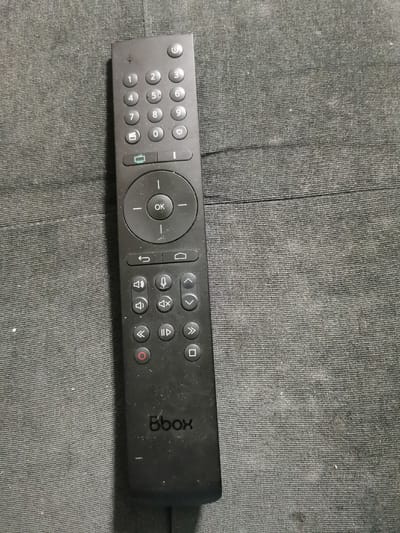 A black remote control is lying flat on a dark gray textured surface. The remote has a standard layout with a power button at the top, followed by a numeric keypad (1-9, 0), and a heart-shaped button. Below the keypad are two small buttons, one with a television icon and the other with a vertical line icon. A circular navigation pad with an "OK" button in the center is below these. The navigation pad is surrounded by directional arrows. Beneath the navigation pad are playback controls (rewind, play/pause, fast forward), a record button, and a square button. A microphone icon is also present on the remote. The brand name "Bbox" is visible at the bottom of the remote. The textured surface appears to be a fabric, possibly a couch or a carpet. There are no people or any other objects visible in the image. The lighting is overhead, casting subtle shadows. There is no indication of the location within the city of Bourg-lès-Valence, France, in the provided image.