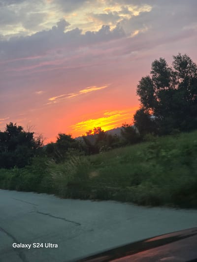 The image depicts a sunset scene over a rural landscape, presumably in Gorna Rositsa, Bulgaria. The sky dominates the upper two-thirds, exhibiting a vibrant color gradient. At the horizon, a brilliant yellow-orange glow emanates from the obscured sun, transitioning upwards into shades of deep orange, then vibrant pinks, purples, and finally pale grays and blues at the very top. Scattered clouds, some tinged with bright yellow and orange from the direct sunlight, and others appearing darker gray and purple, are visible across the sky. Several long, thin, pinkish-purple cloud streaks run horizontally through the mid-sky.
The middle ground features a silhouette of dense vegetation and rolling hills against the bright sunset. Dark green trees and shrubs, including a prominent cluster on the left and a continuous line across the horizon, obscure the distant terrain. A bare, branching tree or shrub stands out as a dark silhouette against the fiery sky in the left-center.
The foreground, occupying the bottom third of the image, shows a stretch of paved road (likely asphalt or concrete) on the left, displaying cracks and an uneven surface. Adjacent to the road and extending across the right side of the foreground is an area of varied green foliage, including tall grasses and bushes, some appearing slightly blurred, suggesting motion. A dark, reflective surface, possibly part of a car's interior, is visible in the bottom right corner, reflecting a reddish hue from the sunset. The text "Galaxy S24 Ultra" is watermarked in white in the bottom left corner of the image, superimposed on the road surface. No people or animals are visible.
