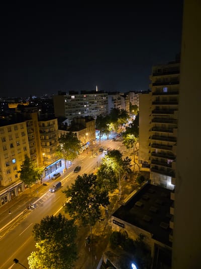 This is a high-angle, nighttime photograph of a street scene in Montrouge, France. The image captures a Parisian-style street lined with multistory apartment buildings. A few cars and a van are visible on the street, and streetlights illuminate the scene, casting yellow light on the wet pavement, suggesting recent rain or mist. Trees with dense foliage are interspersed along the sidewalks, their leaves lit by the streetlights. In the distance, the Eiffel Tower is faintly visible, a common landmark in the Parisian skyline. The perspective suggests the photo was taken from a balcony or window of a tall building overlooking the street and surrounding architecture.