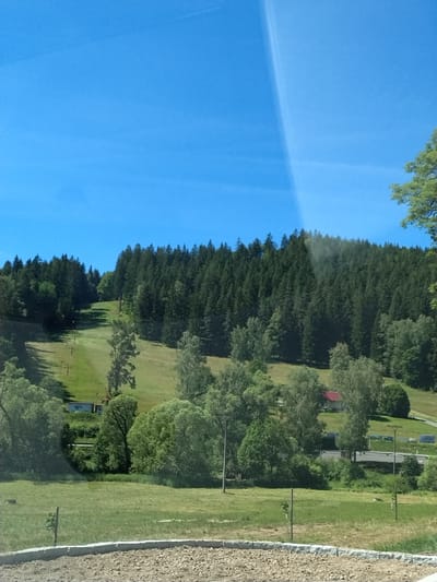 The image depicts a hillside with a ski lift ascending a grassy slope. The slope is bordered by a dense forest of evergreen trees that covers the top of the hill. In the foreground, there is a grassy field with scattered trees and a gravelly area. A small, red-roofed building is visible amidst the trees on the hillside. A road with power lines and signs runs across the middle ground. The sky is clear blue with a faint, bright streak of light. The scene appears to be a natural landscape, possibly a recreational area, given the presence of the ski lift.