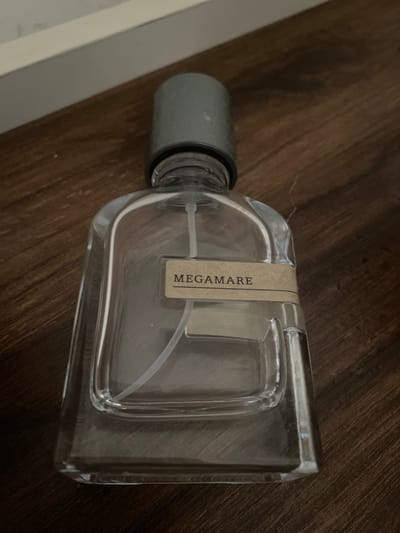 The image shows an empty glass perfume bottle lying on a wooden surface. The bottle has a silver cap and a light brown label with the text "MEGAMARE" printed vertically in black lettering. The perfume bottle is transparent, and a clear tube for spraying is visible inside. The wooden surface has a dark brown, grainy texture, suggesting it might be a table or a shelf. The lighting is dim, casting subtle shadows, but there are no strong indications of the time of day or weather. The scene appears to be an indoor setting, likely a home or a retail environment, given the presence of the perfume bottle and the wooden surface. No people are visible in the image. The overall mood is still and objects are centered.