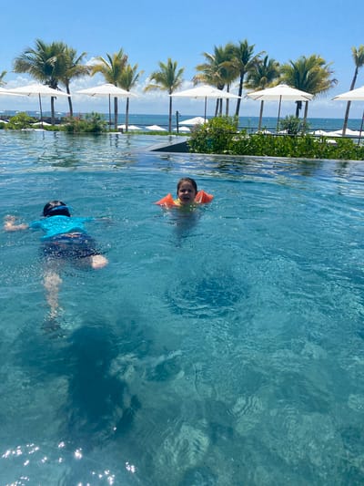 Two children are swimming in a pool. One child is wearing orange arm floaties and is visible from the chest up. The other child is underwater wearing blue swimwear and goggles. In the background, there are palm trees, white umbrellas, green vegetation, and a blue sky. The location is Marina Maroma, Mexico.