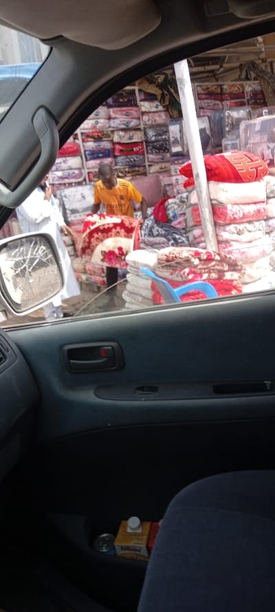 The image depicts an outdoor market scene in Kofar Ruwa, Nigeria, viewed from the interior of a vehicle. The foreground shows the car's dark blue/grey dashboard, door panel with a black handle featuring a red lock indicator, and a storage compartment holding a yellow "CHIVITA" juice carton and a silver can. A dark blue/grey car seat is visible in the lower right. A severely cracked side mirror reflects a distorted portion of the scene.
Outside the vehicle, the primary subjects are two males and numerous textile goods. A dark-skinned male in a yellow short-sleeved shirt, bearing "Emirates Fly Better" text, is positioned in the midground, bent forward, handling a red and cream patterned blanket with floral designs. To his left, another dark-skinned male, partially obscured, wears a long white robe and has his right arm raised, appearing to be speaking or gesturing.
The background and midground are dominated by stacks of folded blankets or duvets, many encased in clear plastic packaging, displaying various patterns and colors. These goods are arranged on shelves against a wall and in large piles on what appear to be display tables or the ground. A prominent stack of red blankets with a white pattern is visible to the right. A white vertical pole, likely a structural element, stands near the center-right. The scene indicates a commercial setting, possibly a textile or bedding stall.