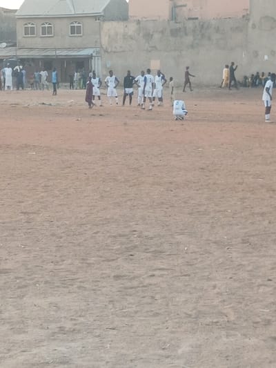 This image, captured in Yalwa, Nigeria, depicts an outdoor community football (soccer) match or practice. The scene is set on a vast, unpaved sandy or dirt field that occupies most of the frame, characteristic of a local sports ground.
In the lower left section, several buildings, including a multi-story structure with distinct windows, are visible, indicating a semi-urban environment. Closer to these buildings, a group of people, presumably football players, are gathered on the field. They are predominantly dressed in white and some in blue jerseys. One player, wearing a white jersey with the number '8' clearly visible on their back, stands further out on the field. Another player in a white uniform is seen lying on the ground, possibly resting or having fallen during play. Along the edge of the field near the buildings, a crowd of onlookers observes the event. These spectators wear a mix of casual attire and traditional West African garments.
The lighting suggests it is daytime, likely either morning or late afternoon, with soft, diffused light and no harsh shadows. The weather appears clear. While specific emotions are not discernible due to the distance and image quality, the gathering signifies a lively community engagement centered around sports. The informal nature of the field and the diverse crowd highlight a local sporting event typical of the region.