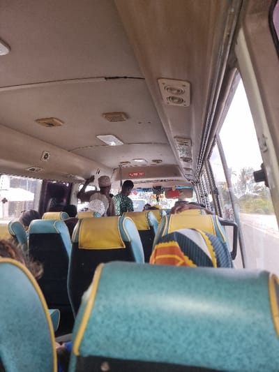 The image displays the interior of a public transit minibus, likely a 'dala-dala', situated in Paje, Tanzania. The perspective is from a passenger seat towards the front of the vehicle.

The bus interior features rows of passenger seats upholstered in teal-blue fabric with yellow headrest covers and trim. The ceiling is light-colored, exhibiting a linear crack or seam, and contains multiple recessed rectangular air vents and overhead lights.

Several individuals are present within the vehicle. Two adult males stand in the central aisle towards the front. One wears a light-colored head covering and shirt. The other wears a green patterned shirt. Other passengers are seated, visible as partial figures or from the back of their heads, distributed throughout the seating area. A prominent patterned headrest cover is visible on the seat directly in front of the viewer's perspective.

Through the large windows on the right side of the bus, an exterior scene is visible. A paved road runs parallel to the vehicle, bordered by green foliage and trees under a bright, overcast or light sky. The vehicle appears to be in operation on a roadway within Paje.
