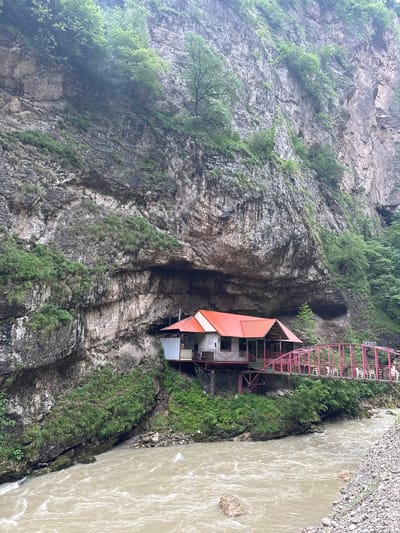 The image captures a scenic view of a building situated beneath a massive, rugged cliff face, adjacent to a flowing river. The building, with its striking red roof, is perched on stilts and connected to a pink metal bridge spanning the river. The bridge has white chairs set up on it, suggesting it might be an outdoor seating area. The cliff behind the building is heavily textured, with visible rock formations and patches of green vegetation clinging to its surface. The river itself appears to be muddy and fast-flowing, with several rocks visible in its current. Lush green trees and foliage are abundant, both on the cliff face and along the riverbanks, indicating a natural, possibly mountainous, environment. A sign on the bridge reads "КАФЕ НА МОСТУ," which translates to "Cafe on the bridge." This suggests the building is a cafe or restaurant, and its location is in Khushto-Syrt, Russia.