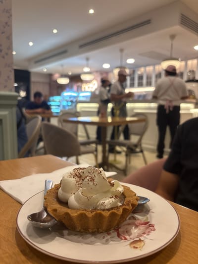 A dessert, possibly a tart with cream, sits on a floral-patterned plate with a spoon on a wooden table. The background shows a blurred interior of a restaurant, with people and tables visible. The location is Veracruz, Mexico.